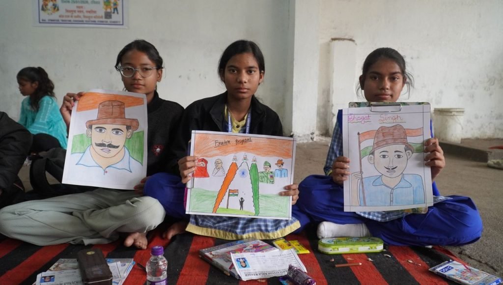 Three children hold patriotic drawings highlighting Indian freedom fighters in a classroom setting.