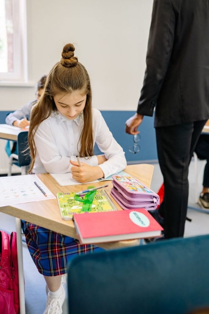 A young girl sits at a desk writing attentively in a classroom setting.